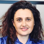 A woman with wavy brown hair in a blue blouse smiles at the camera.