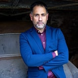 Man with short hair and beard in a blue blazer and maroon shirt stands with arms folded in front of a medbridge-style background.