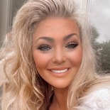A person with wavy blonde hair smiles in front of a window, reflecting on Medbridge's digital healthcare impact.