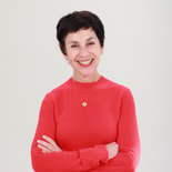 A woman with short dark hair smiles at the camera, wearing a red sweater and a necklace, in front of a plain white background.