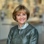 A smiling person with short hair, in a leather jacket and patterned scarf, stands inside.