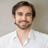 A man with medium-length dark hair and a beard, wearing a white shirt, smiles confidently at the camera, embodying digital healthcare innovation for medbridge.