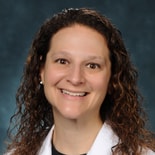 A person with curly brown hair smiles at the camera in a white coat and black shirt, set against a textured blue-green background for Medbridge.