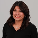 A person with dark hair smiles, wearing a black top and jewelry, against a neutral background.