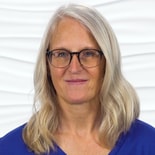 Someone with long gray hair and glasses, in a blue shirt, is in front of a white textured background.