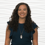 A woman with curly hair in a blue top smiles against a white background, wearing a pendant necklace.