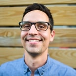 Person with glasses and short hair in a blue shirt stands against a wooden background.