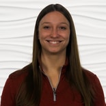 A smiling person with long brown hair wears a red jacket against a white, wavy background for medbridge.