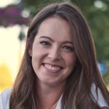 A smiling woman with long brown hair, in a white shirt, stands against blurred greenery, representing medbridge's harmony of nature and technology.