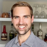 Bearded man in a checkered shirt stands smiling before shelves of bottles and glassware.
