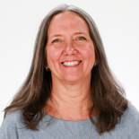 A smiling woman with long gray hair in a gray top poses against a white background, embodying the compassionate spirit of digital healthcare and medbridge innovations.