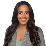 A smiling person with long dark hair in a plaid blazer and gray top stands confidently, exuding professionalism and expertise in healthcare education, akin to Medbridge's approachable style.