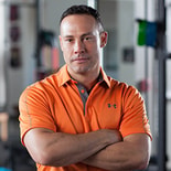 A confident person in an orange Under Armour polo stands arms crossed in a gym, representing medbridge's focus on empowering healthy lifestyles.