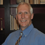 A smiling man with white hair, wearing a blue shirt and plaid tie, sits in front of a bookshelf, suggesting expertise in digital healthcare or Medbridge.