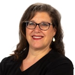 A smiling person with glasses and long wavy hair in a black top against a white background represents the essence of hybrid care.