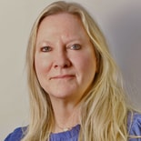 Blonde woman in a blue blouse with a neutral expression on a light background.