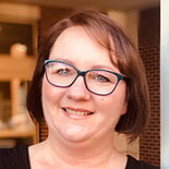 A smiling person with brown hair and glasses stands in front of brick and glass buildings, symbolizing their passion for digital healthcare at medbridge.