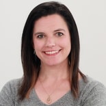 A person with long, dark brown hair in a grey top smiles at the camera.