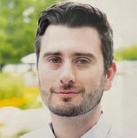 A bearded man with short dark hair smiles slightly at the camera against a blurred backdrop of greenery and flowers.