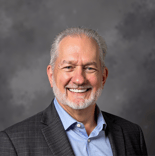 Smiling older man with gray hair and beard in a blue shirt and plaid blazer stands before a gray background for medbridge.