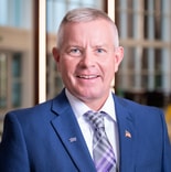 A smiling man with short gray hair in a blue suit and plaid tie stands indoors, wearing two lapel pins, at medbridge.