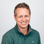 A man with short curly hair in a green zip-up sweater smiles at the camera, representing digital healthcare with medbridge.