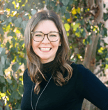 A smiling woman with long brown hair and glasses, in a black top, stands outdoors on a sunny day, reflecting her dedication to healthcare education at medbridge.