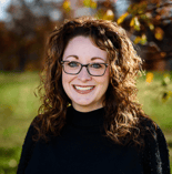 A woman with curly brown hair and glasses smiles outdoors, framed by blurred autumn leaves, showing warmth seen in medbridge settings.