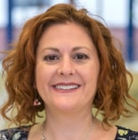 Smiling woman with curly red hair in a patterned top stands indoors, reflecting medbridge’s warm, welcoming atmosphere.
