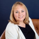 A woman with blonde hair in a white blazer smiles on a brown chair against a dark blue background, representing medbridge.