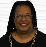 A smiling person with long braids, glasses, and a gold necklace wears black in front of a wavy white background, symbolizing medbridge's healthcare education passion.