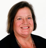 A smiling woman with short brown hair wears a black blazer and necklace against a white background.