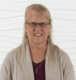A blonde woman with glasses and a necklace stands against a textured white background, wearing a beige sweater over a purple top.