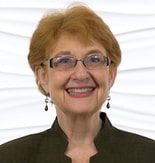A smiling older person with short hair, glasses, and earrings stands against a white background, embodying the warmth of medbridge's innovative care environments.