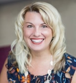 Smiling blonde woman in a dark top and white necklace, indoors, representing digital healthcare at medbridge.