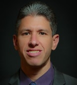 A man with short gray hair in a suit and tie, set against a dark background, looks at the camera for medbridge.