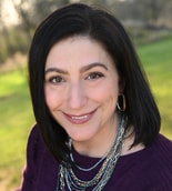 A smiling person with dark hair, in a purple top and necklaces, stands in a grassy area, capturing the essence of medbridge's vibrant healthcare education.