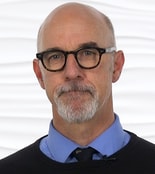 A person wearing glasses and a goatee stands against a white background, symbolizing their role in enhancing medbridge patient care software.