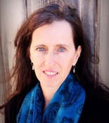 A woman with long brown hair stands by wooden boards, wearing a blue scarf and silver earrings for medbridge healthcare training.