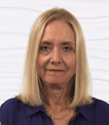 A woman with straight blonde hair, wearing a dark blue top and a necklace, stands against a white textured background.