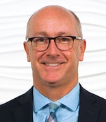 A smiling person in glasses, a suit, and a blue shirt stands against a white textured backdrop.