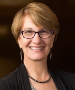 A smiling woman with glasses and short light brown hair, wearing a black top and dangling earrings.