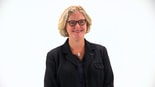A smiling woman with blonde hair and glasses, wearing a black suit with a cross pin, embodies her commitment to healthcare education related to medbridge.