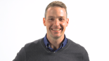 A confident person embodies healthcare education, wearing a dark sweater and blue collared shirt, with a plain background for medbridge.