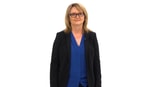 A woman in glasses, a black blazer, and a blue blouse stands against a white background.