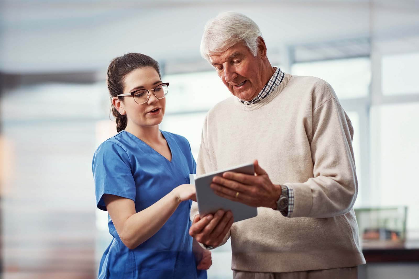 A nurse in blue scrubs talks with an older man in a beige sweater about a clipboard, representing Medbridge's commitment to quality care.