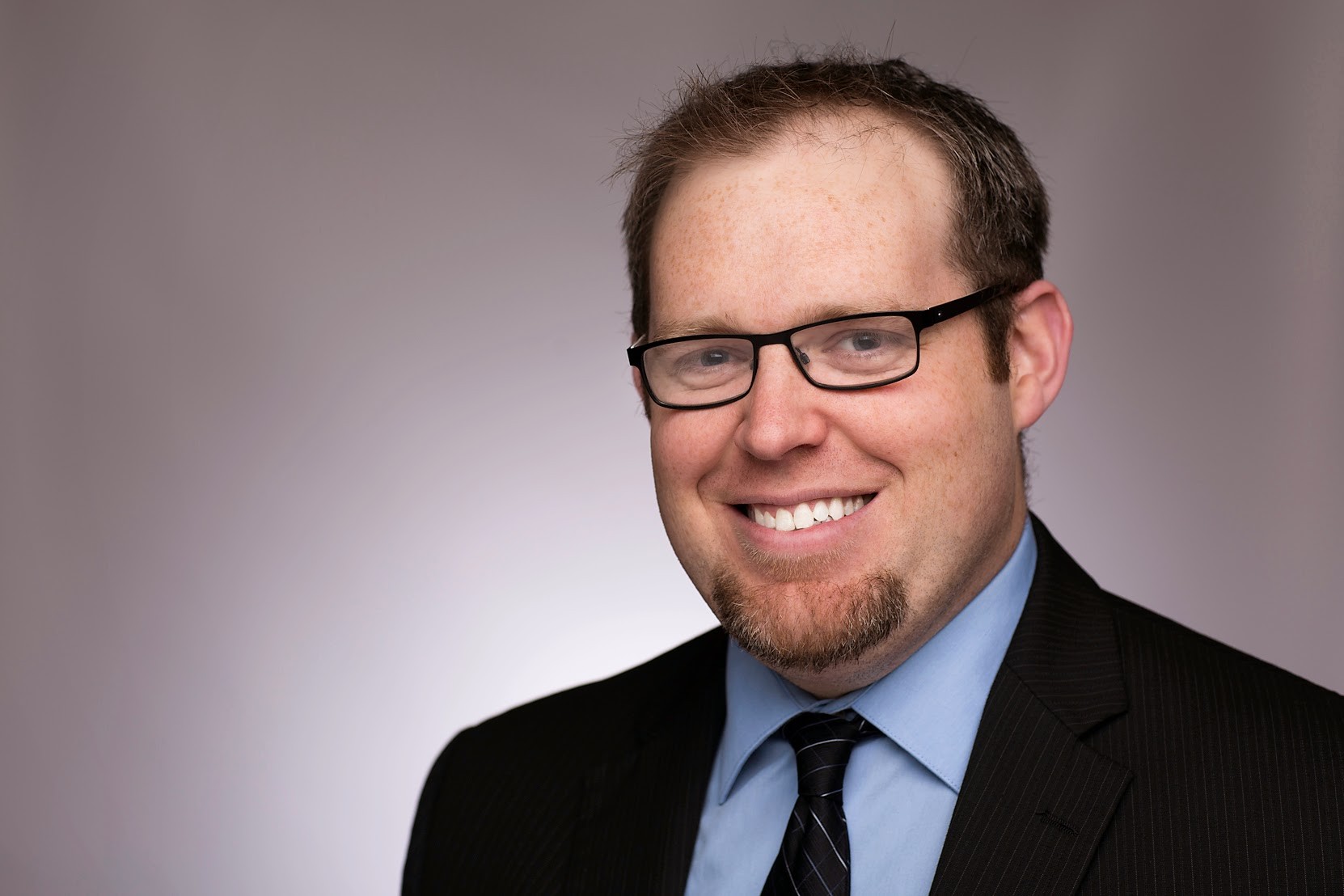 Smiling man in a suit, glasses, and tie in a portrait against a plain background for Medbridge.