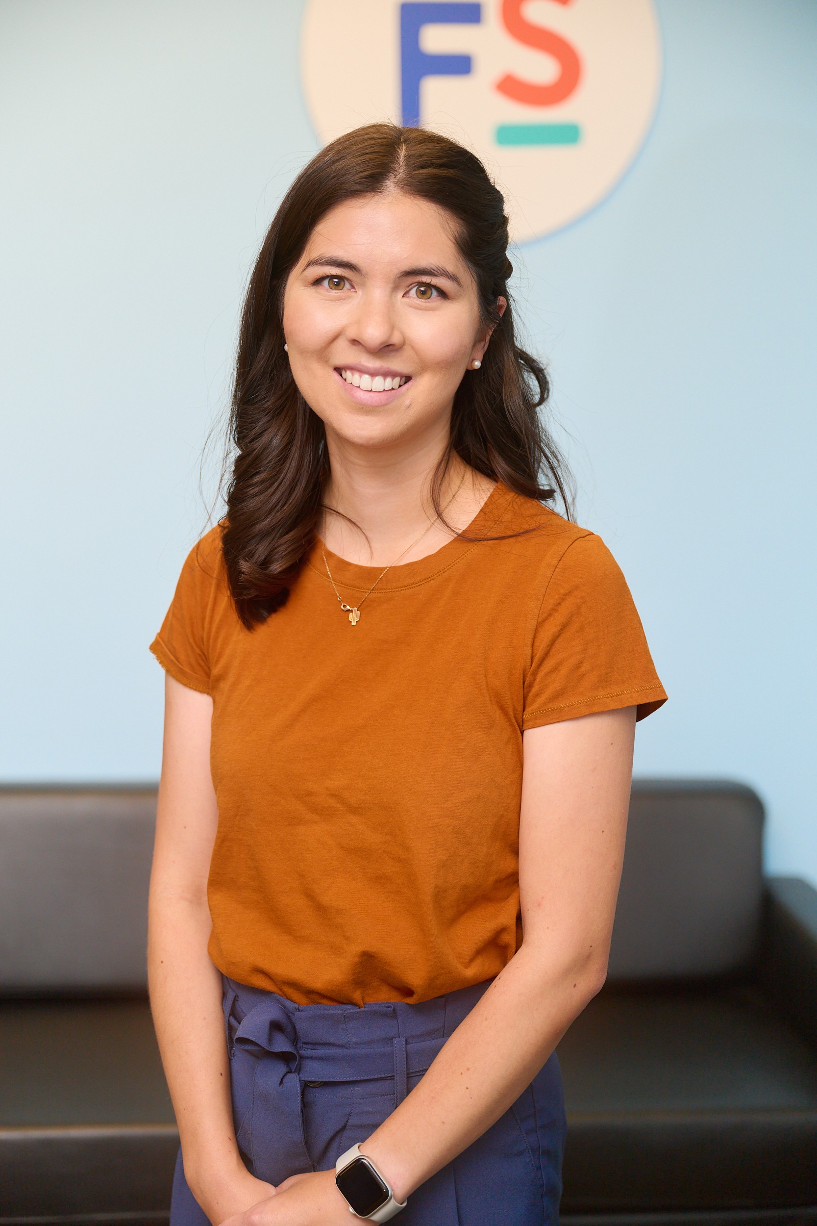 Smiling woman in orange shirt stands by black couch and medbridge logo wall indoors.