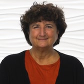 A smiling person with curly brown hair wears an orange top and black cardigan in front of a textured white background.