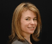 A woman with shoulder-length blond hair in a gray jacket smiles slightly at the camera against a black background.